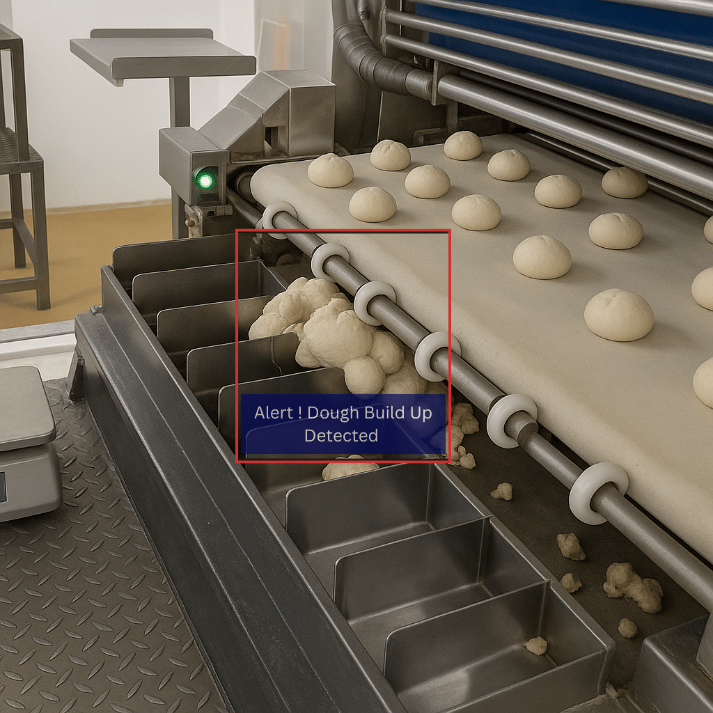 An industrial bakery conveyor belt with rows of dough balls. A build-up of dough is visible accumulating below the rollers and spilling into metal collection trays, indicating excess dough overflow.