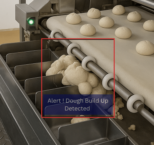 An industrial bakery conveyor belt with rows of dough balls. A build-up of dough is visible accumulating below the rollers and spilling into metal collection trays, indicating excess dough overflow.